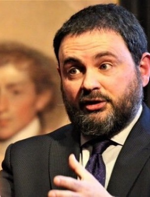 Ben Railton. White man in a suit and tie with a black beard, mustache, and hair is speaking animatedly at an academic talk