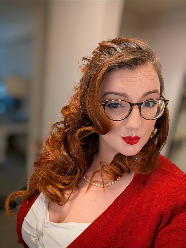 Vaughn Joy. White woman with red curled hair, glasses, red lipstick, a white top, red cardigan, and pearls looking at the camera